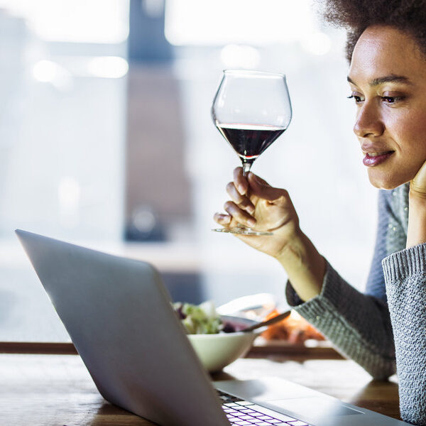 Smiling black woman drinking wine while reading online recipes in the kitchen. Young African American woman drinking wine while reading recipes over laptop in the kitchen.